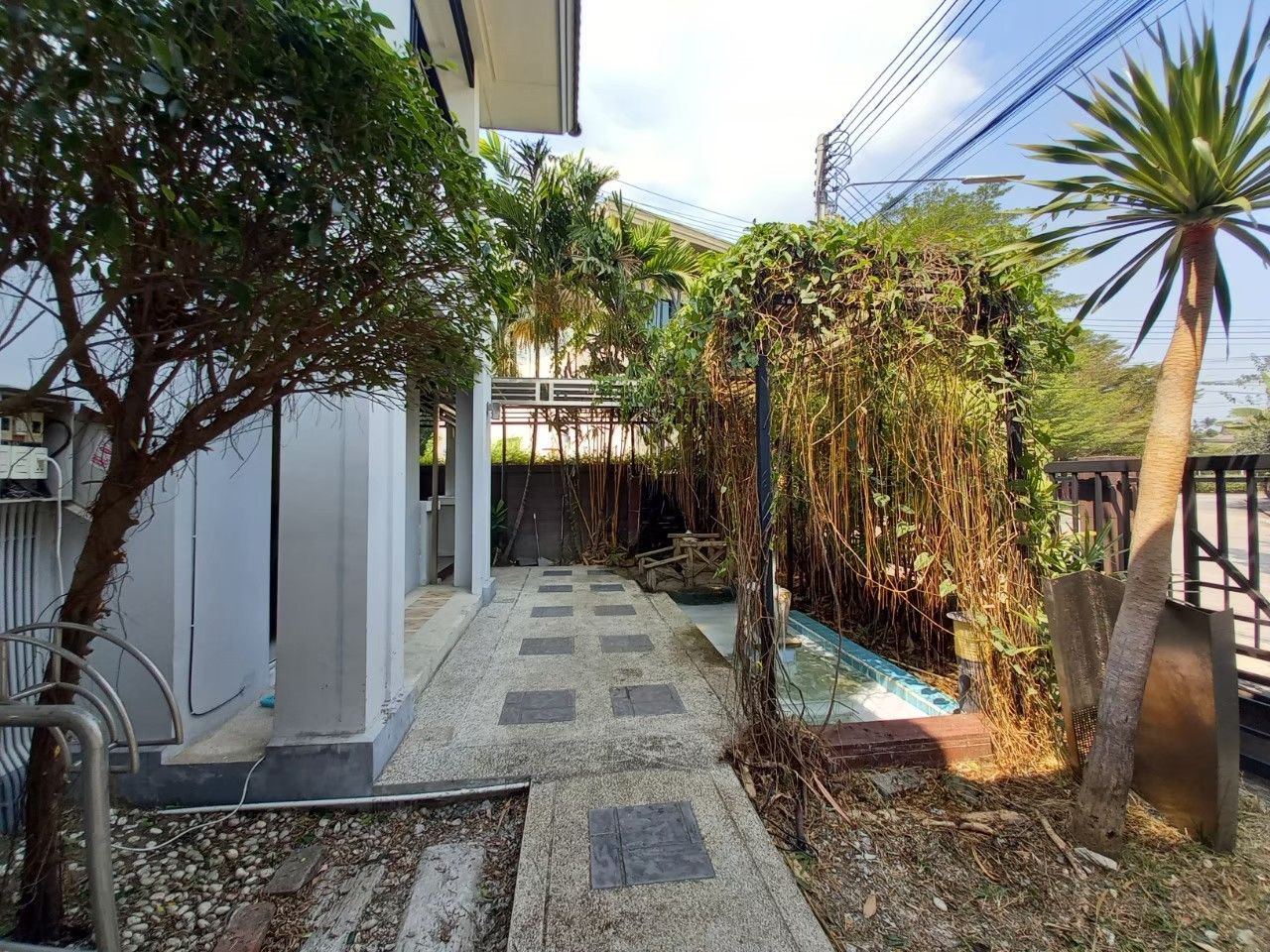 Un estrecho sendero que lleva a un edificio con entrada cubierta, rodeado de exuberante vegetación y una pequeña fuente.