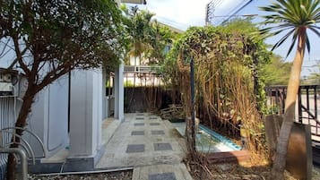 A narrow pathway leading to a building with a covered entrance, surrounded by lush greenery and a small water feature.