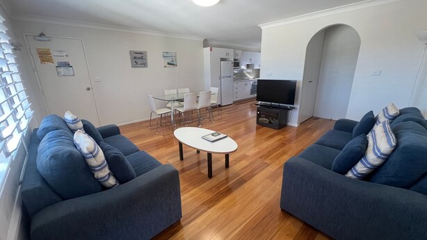 Una sala de estar moderna con dos sofás, una mesa de centro y un televisor. Se observa un comedor con sillas y una cocina al fondo.