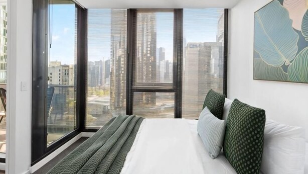 A bedroom with a large bed, a city view through floor-to-ceiling windows, and a cityscape visible outside. - A Modern 2BR Apt with City Views, FREE Parking (Southbank)