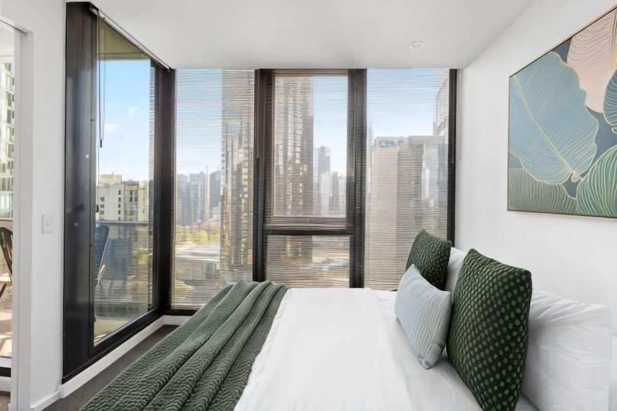 A bedroom with a large bed, a city view through floor-to-ceiling windows, and a cityscape visible outside.