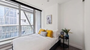 Een slaapkamer met een groot raam, een bed met witte lakens, een nachtkastje met een lamp, en een lijst met een afbeelding aan de muur.