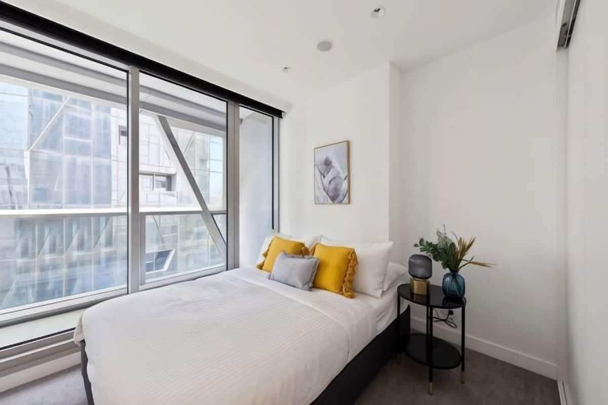 A bedroom with a large window, a bed with white bedding, a bedside table with a lamp, and a framed picture on the wall.
