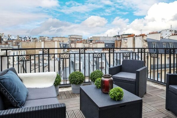 A rooftop terrace with wicker furniture, potted plants, and a city view.