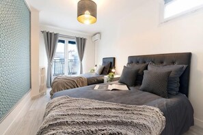 A bedroom with a large bed, a bedside table with a lamp, a window with curtains, and a balcony.