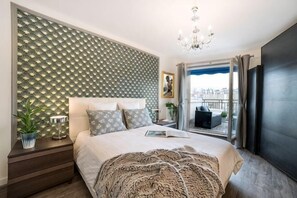 A bedroom with a patterned wall, a bed with pillows, a bedside table with a lamp, and a view of the city through a window.