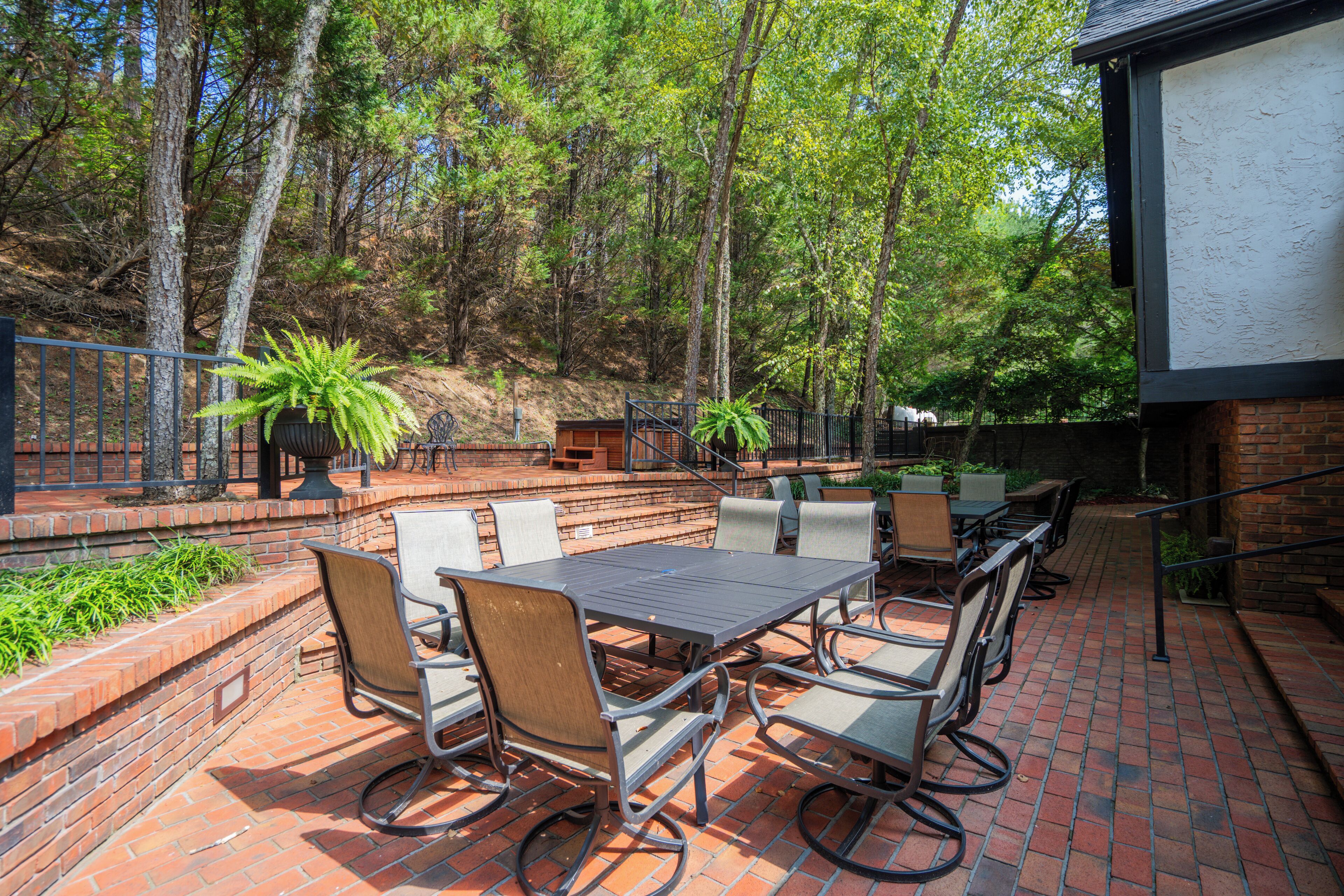 An outdoor patio with a brick floor, surrounded by trees, featuring a table and chairs, a planter with a fern, and a brick wall.