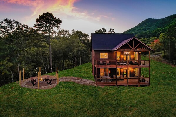 A two-story wooden cabin with a deck, surrounded by trees and a mountain in the background. - 🌅 Sunset Ridge ⛰️ Sunset Mountain Views, Fire Pit, Hot Tub & Game Room (Sevierville)