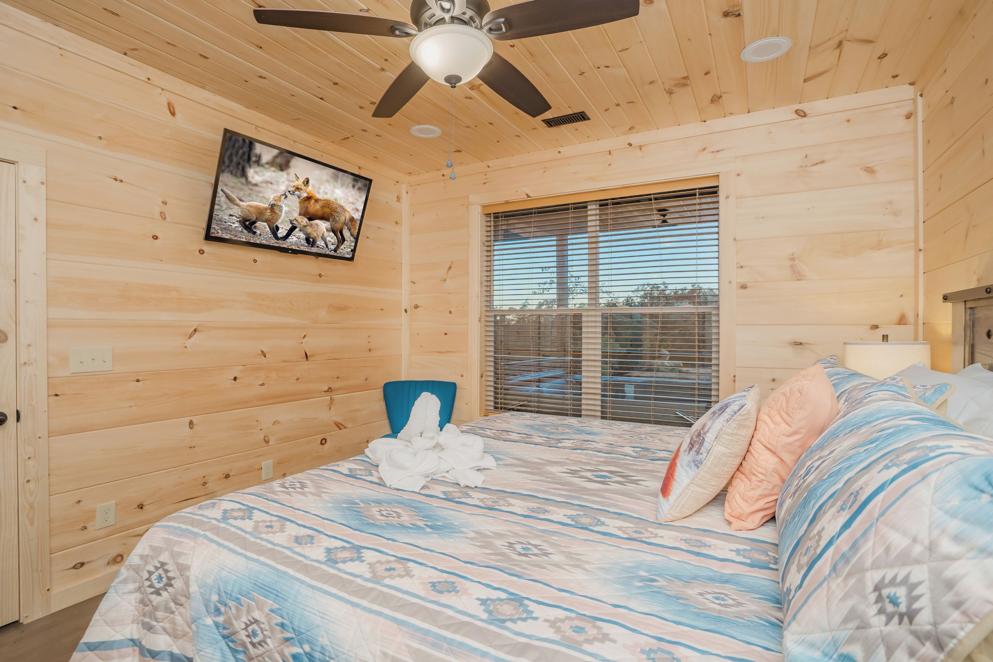 Un dormitorio de cabaña de madera con una cama, un ventilador de techo, una ventana con persianas y un cuadro en la pared.