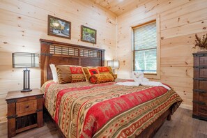 A wooden cabin bedroom with a bed, two wall-mounted lamps, a dresser, and a window with blinds. - 🌅 Sunset Ridge ⛰️ Sunset Mountain Views, Fire Pit, Hot Tub & Game Room (Sevierville)