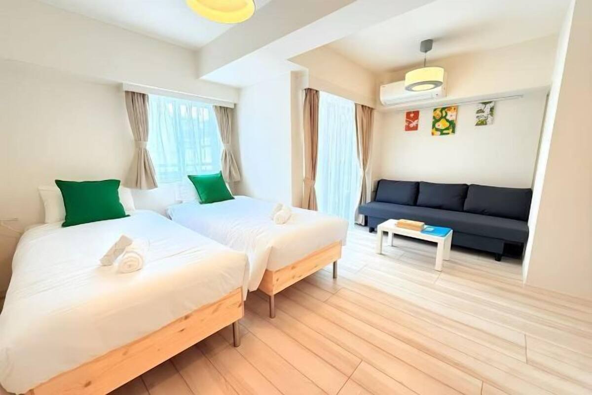 A hotel room with two single beds, a sofa, a small table, and a ceiling fan.