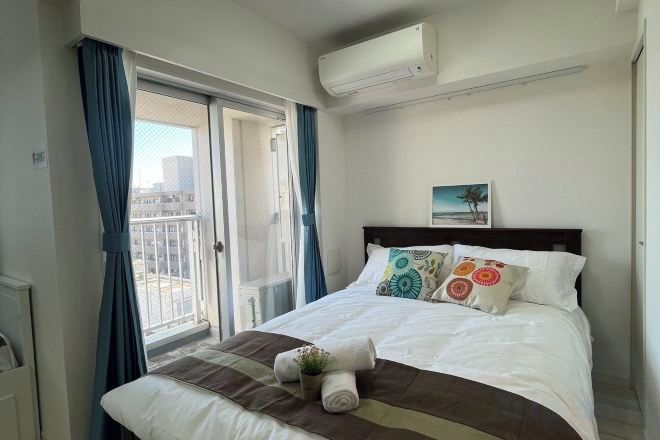 A hotel room with a bed, a window with curtains, an air conditioning unit, and a painting on the wall.