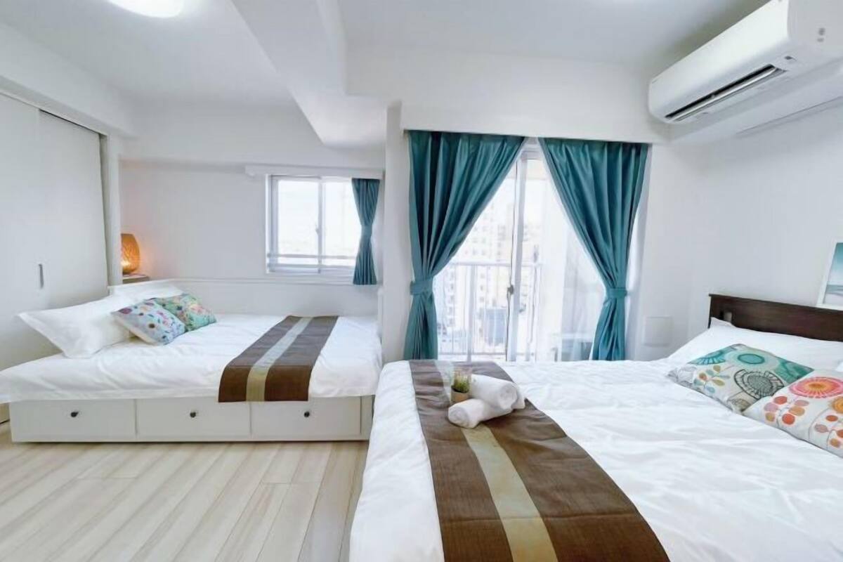 A modern hotel room with two beds, a large window with curtains, an air conditioning unit, and a view of the city.