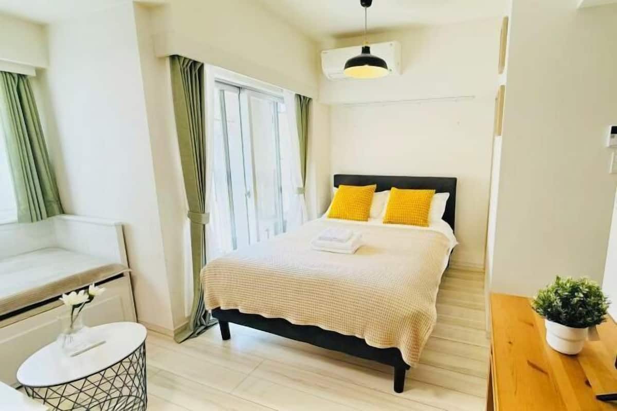 A bedroom with a bed, a bedside table, a window with curtains, and a small potted plant.