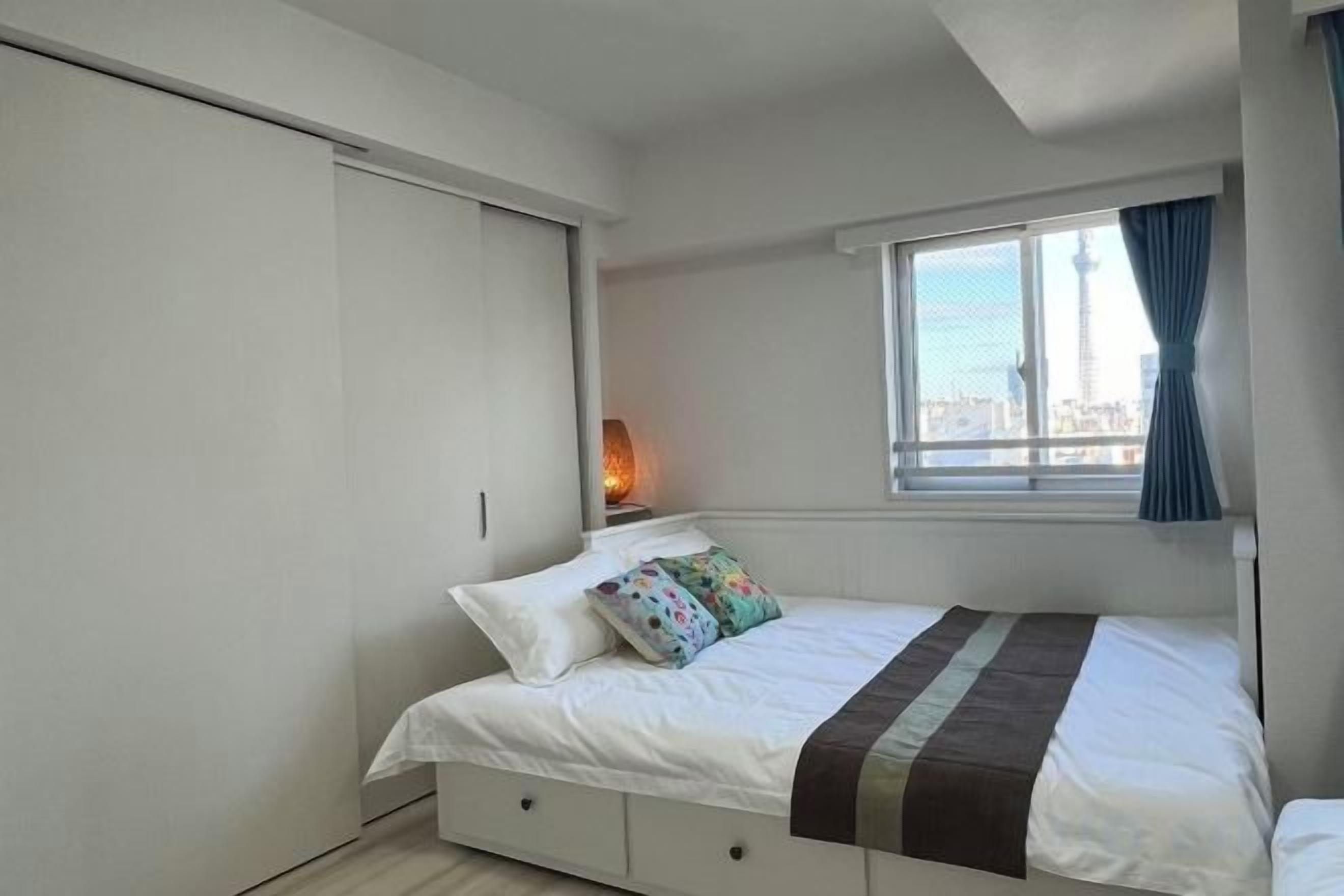 Een slaapkamer met een bed, een raam met gordijnen, een bedlamp en uitzicht op een stadsgezicht.