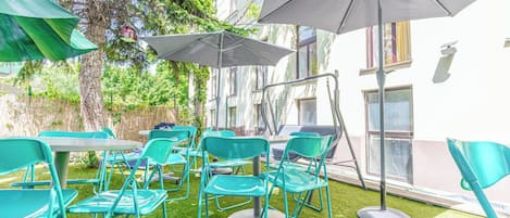An outdoor seating area with green chairs and umbrellas, surrounded by trees and a building.