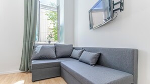 A grey sectional sofa with cushions, a flat-screen TV mounted on the wall, and a window with a view of greenery outside. - 6BR Apt with Garden near Danube (Vienna)