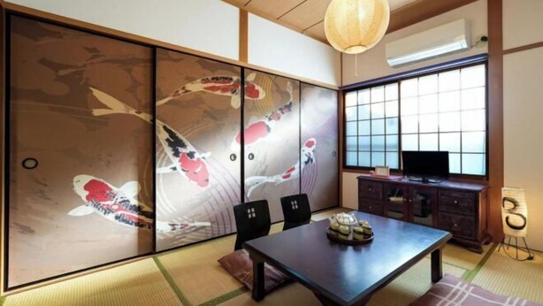 A traditional Japanese room with tatami flooring, a sliding shoji screen with koi fish artwork, a dark wooden table, and a television on a cabinet. - Guesthouse Kinoya (Kyoto)