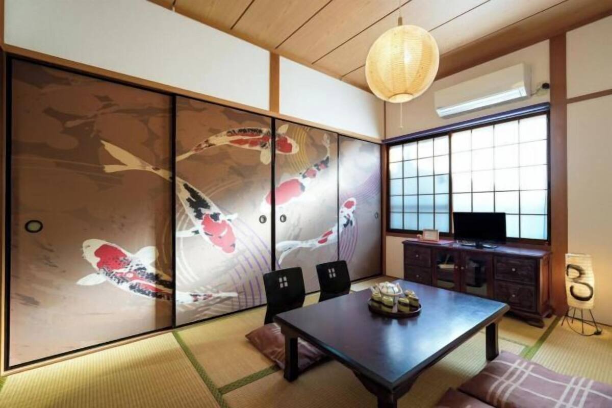 A traditional Japanese room with tatami flooring, a sliding shoji screen with koi fish artwork, a dark wooden table, and a television on a cabinet.