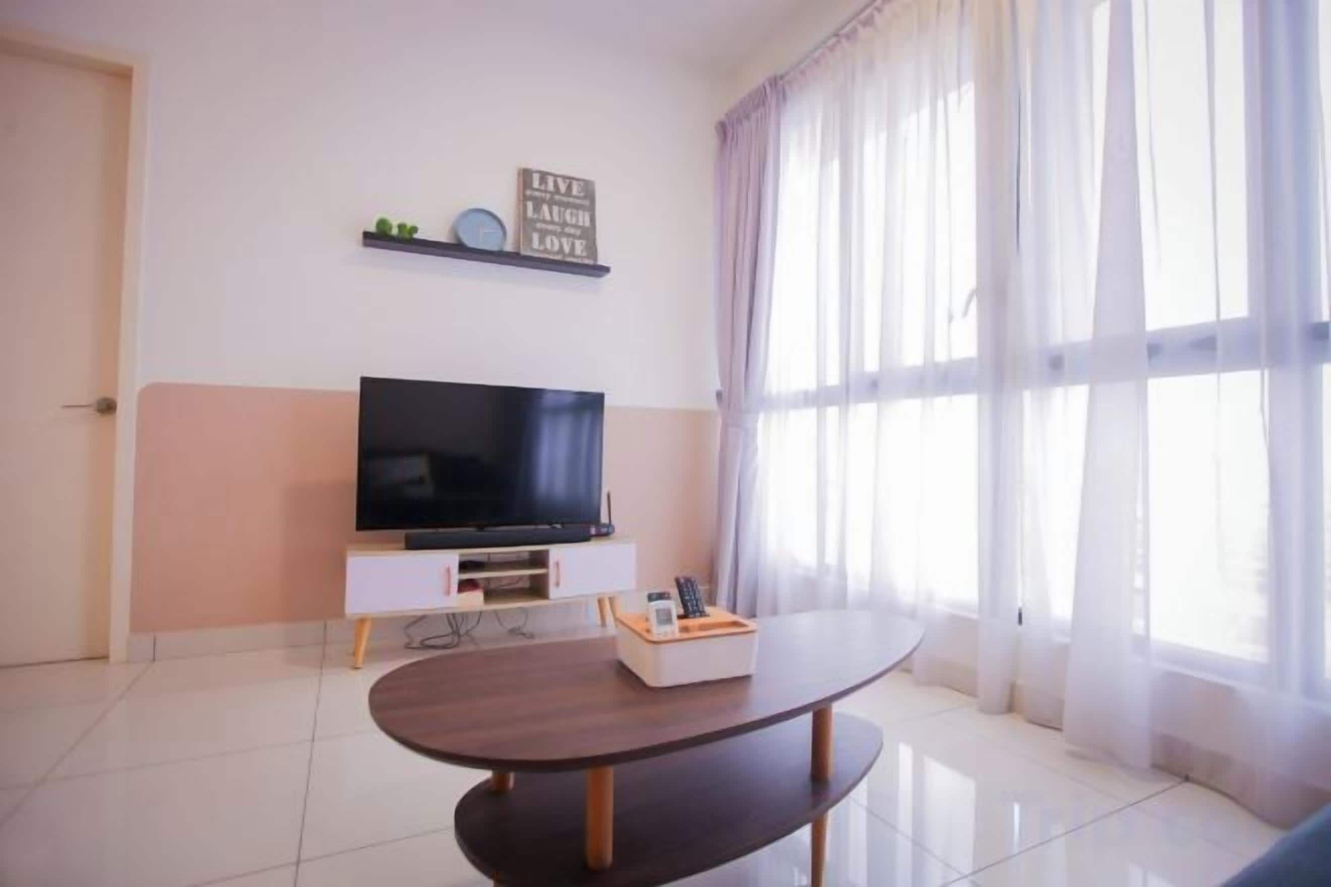 Una sala moderna con un televisor de pantalla plana sobre un mueble blanco, una mesa de centro de madera oscura y un ventanal con cortinas translúcidas.