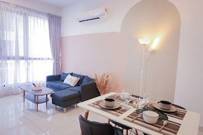 A modern living room with a sofa, dining table, and chairs, air conditioning, and a large window with curtains. - Econest Apartment By The One - Educity Ecobotanic (Gelang Patah)