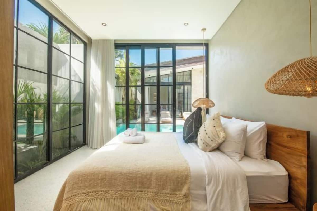 A bedroom with a large window, a bed, a bedside lamp, and a view of a swimming pool.