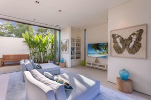 A modern living room with a sofa, a coffee table, and a bookshelf. There is a window with a view of greenery outside. - Sava Villa 7 Villa Aqua (Takua Thung)