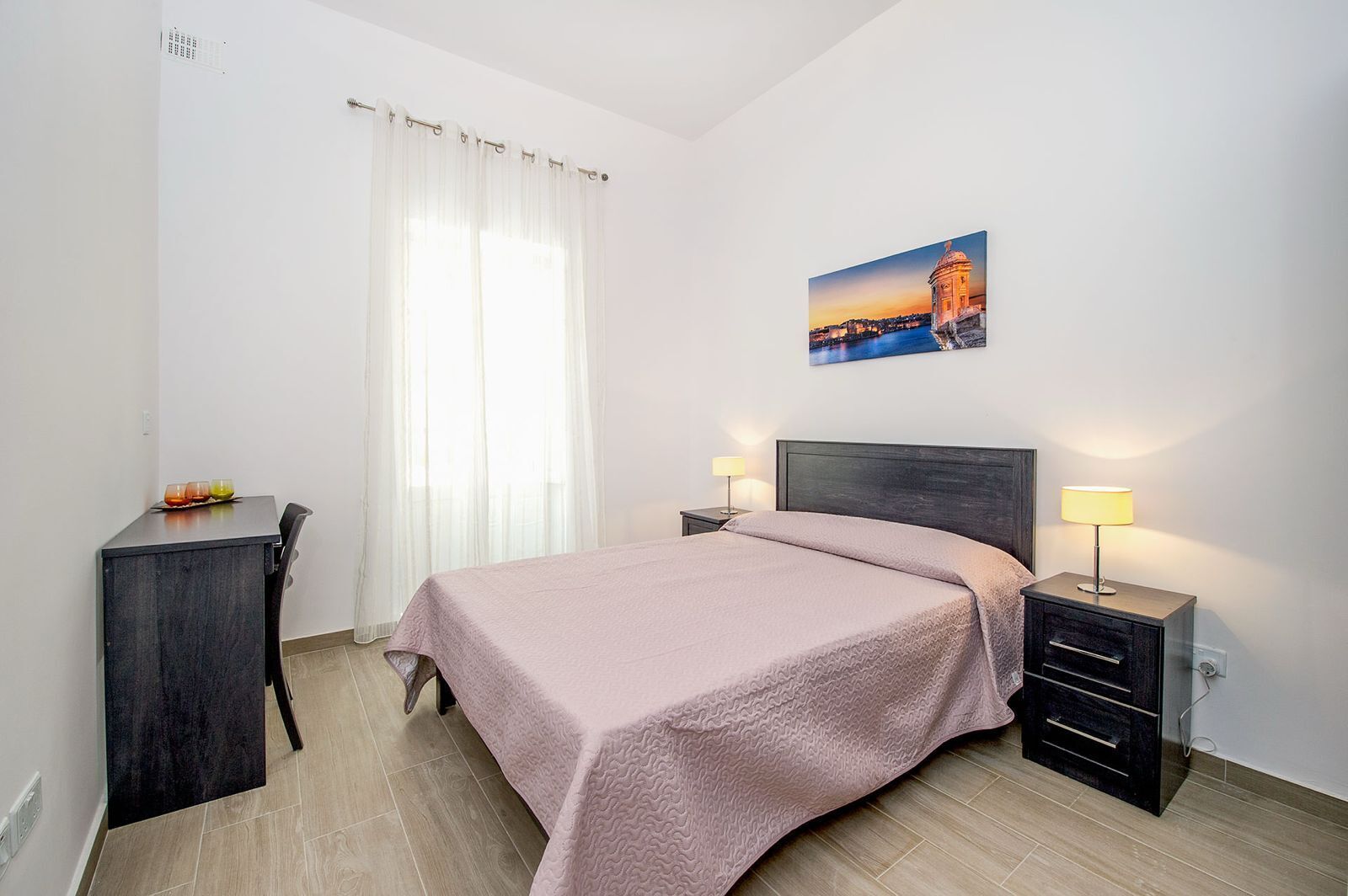 A neatly arranged bedroom with a bed, bedside tables, a desk, and a window with curtains.