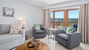 Un salon moderne avec un canapé gris, deux fauteuils, une petite table et un ventilateur de plafond. On aperçoit un balcon avec des rideaux à motifs et une vue sur les bâtiments.