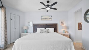 Une chambre à coucher avec un grand lit, un ventilateur de plafond, deux tables de chevet avec des lampes et des œuvres d’art accrochées aux murs.