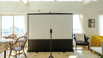 A room with a large window, a tripod-mounted camera, a table with a patterned tablecloth, and a chair.