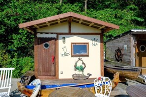 Une petite cabane en bois avec un toit à deux versants, une porte et une fenêtre. On peut voir des bateaux et une planche à pagaie à l’extérieur.