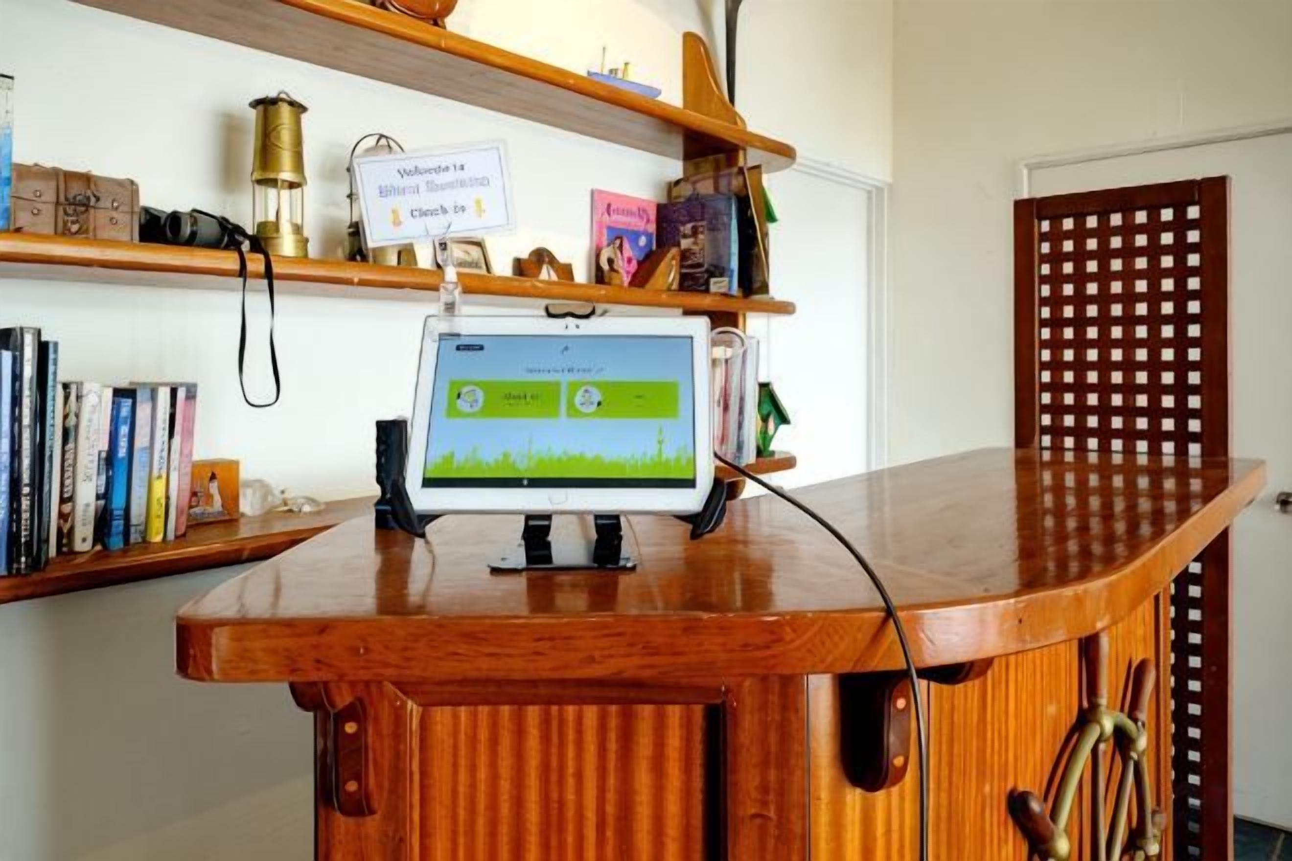 A wooden counter with a computer monitor displaying a green interface, surrounded by books and a sign indicating a waiting area.
