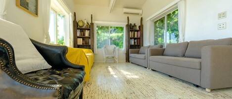 Un salón con un sofá de cuero negro, un sofá gris y una silla tapizada en amarillo. Se observan estanterías y una mesa blanca al fondo.
