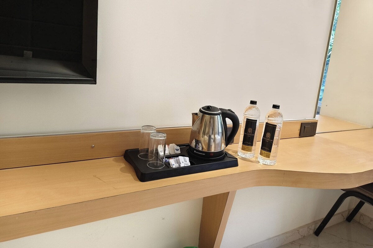 Un escritorio de madera con una bandeja negra que contiene una tetera eléctrica plateada, dos vasos de vidrio y un paquete de cubitos de azúcar. También hay dos botellas de agua y dos botellas de una bebida con etiqueta negra sobre el escritorio.