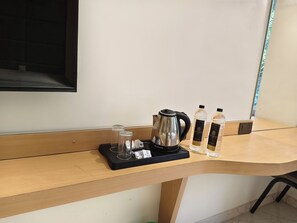 Un escritorio de madera con una bandeja negra que contiene una tetera eléctrica plateada, dos vasos de vidrio y un paquete de cubitos de azúcar. También hay dos botellas de agua y dos botellas de una bebida con etiqueta negra sobre el escritorio.