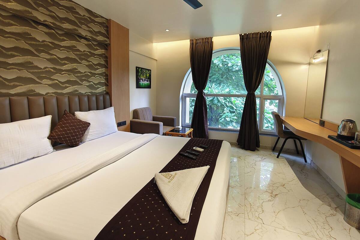 A hotel room with a large bed, a desk, a chair, and a view of greenery through the window.