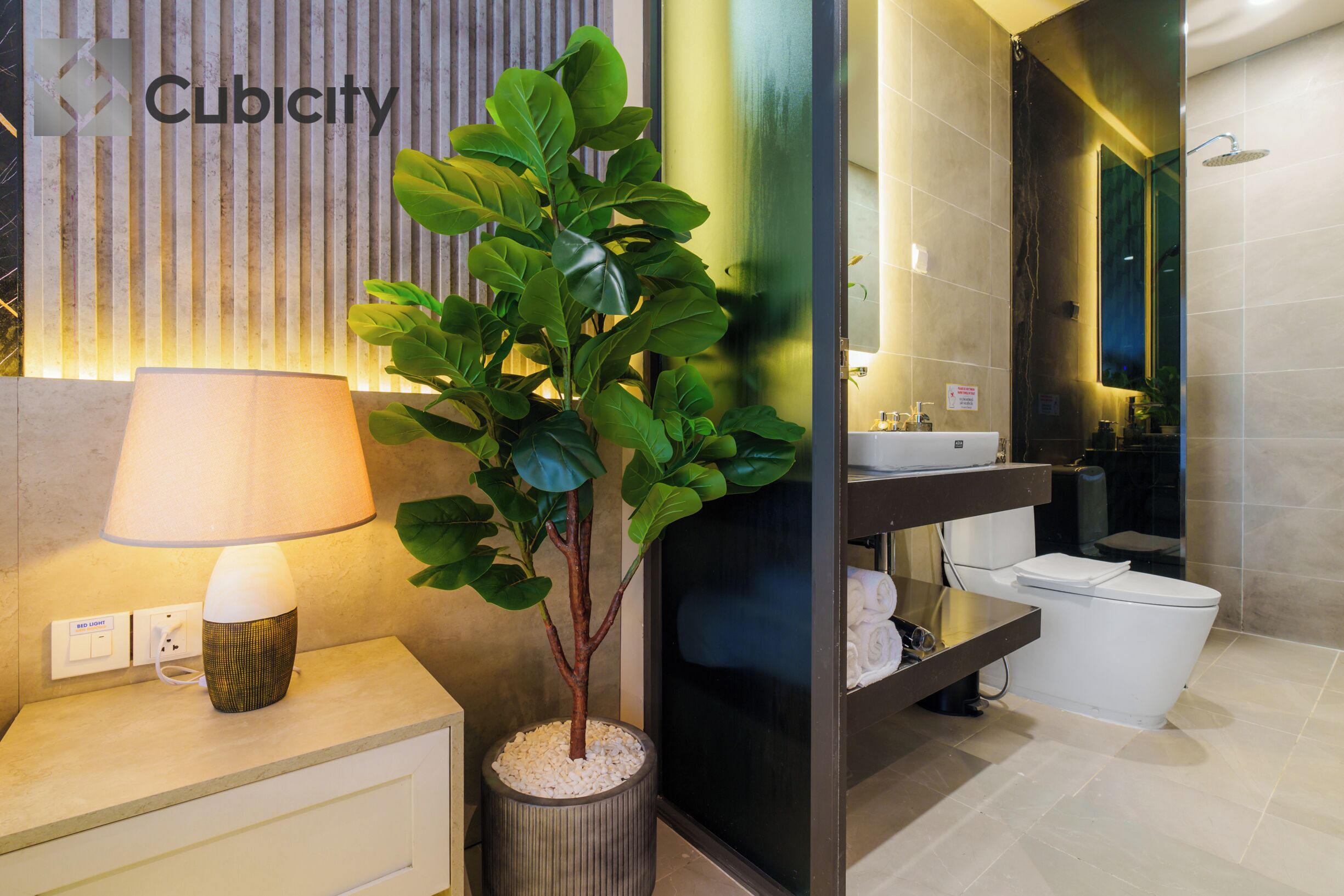 A modern bathroom with a sink, toilet, and a mirror. There is a potted plant and a lamp on a bedside table.