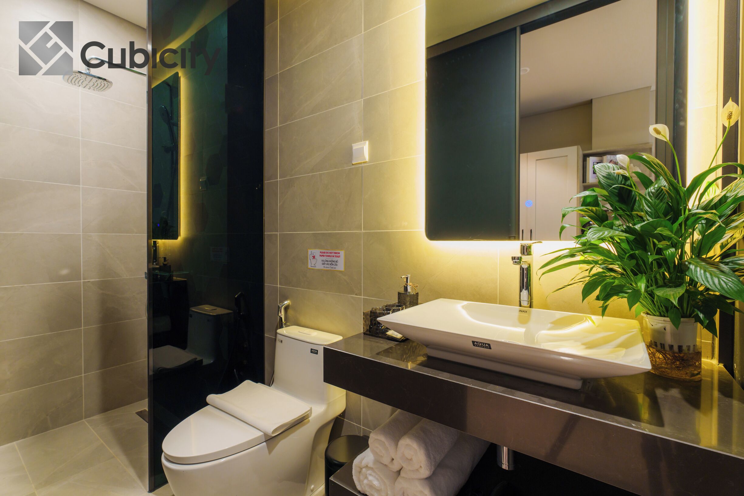 A modern bathroom with a white toilet, a sink, and a mirror. There is a plant on the counter and a towel rack below the sink.