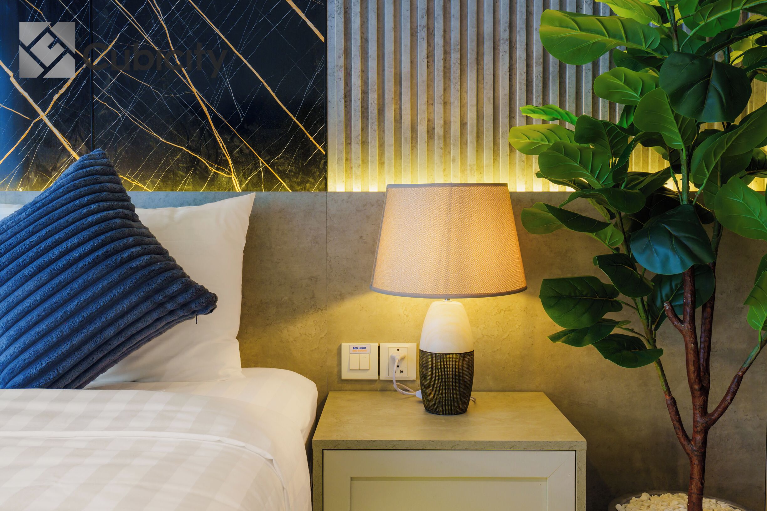 A neatly made bed with a blue pillow, a bedside lamp, and a wall with vertical lines and a plant.