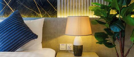 A neatly made bed with a blue pillow, a bedside lamp, and a wall with vertical lines and a plant.