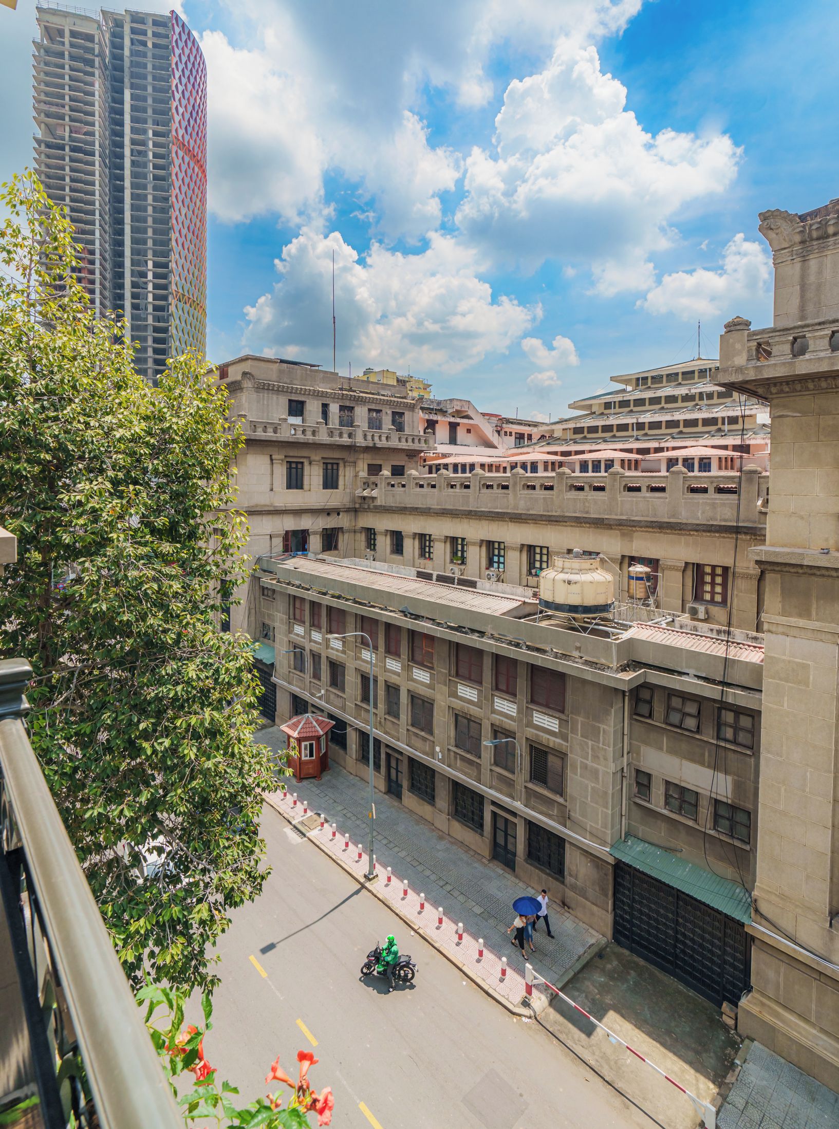 A view of a cityscape with a mix of modern and older buildings, a clear sky, and a street with a few pedestrians and a motorcycle.