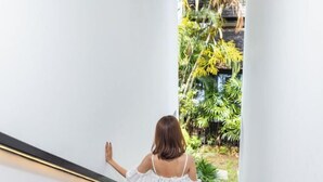 A woman in a white off-shoulder dress standing at the top of a staircase, looking out at a lush garden with various plants and a wooden structure.