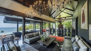 A modern living room with a large sofa, a coffee table, and a chandelier. There is a view of the ocean through large windows.