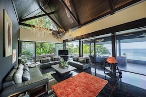 A modern living room with a large sofa, coffee table, and a red rug. There is a television and a chandelier. The room has large windows with a view of the outdoors. - Yin Yangs Villas, Private Oceanfront, Kamala Beach (Phuket)