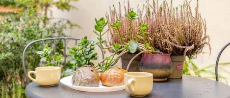 A round metal table with two yellow cups and a plate with two pastries, accompanied by a potted plant with green foliage and a taller plant with brown, spiky leaves.