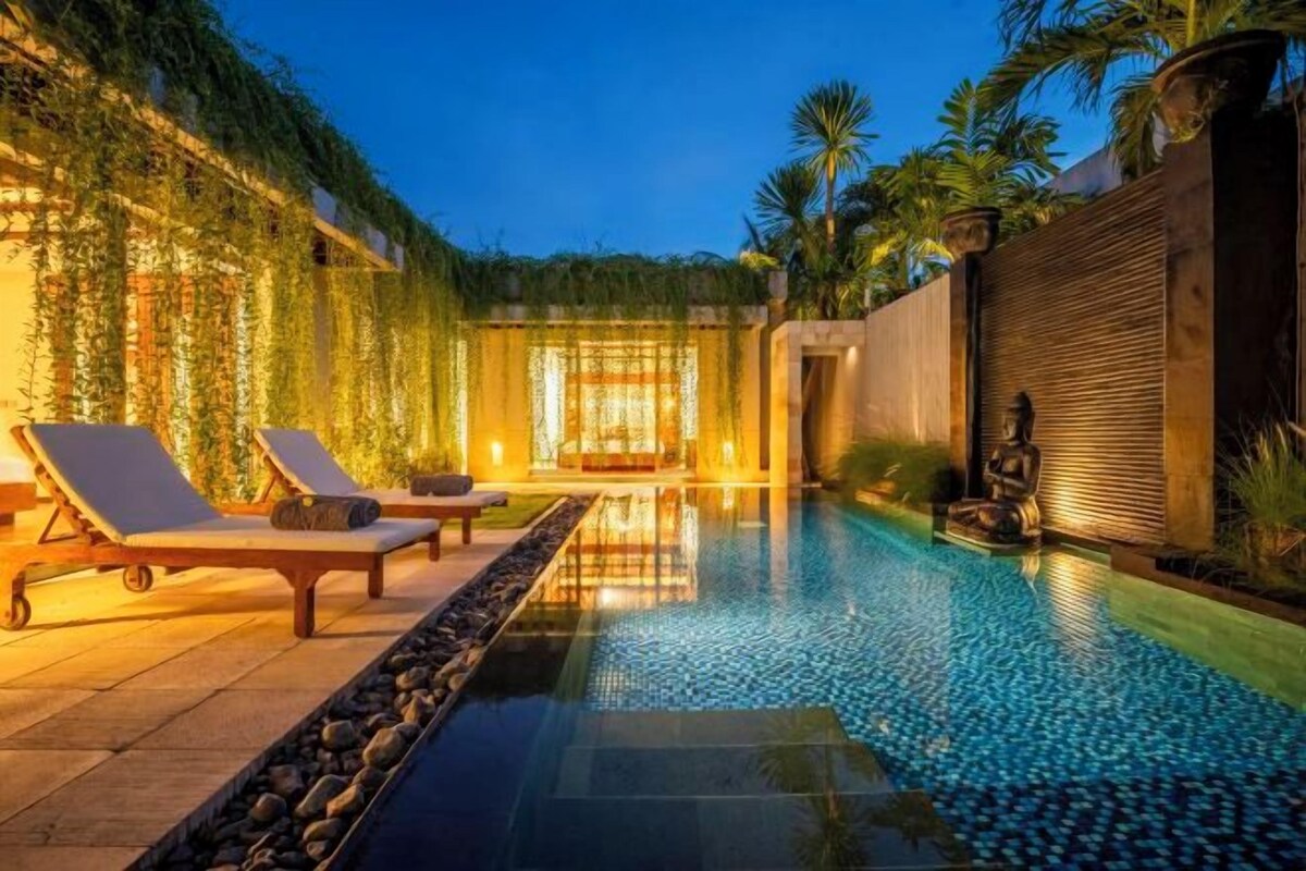 An infinity pool with lounge chairs and a Buddha statue, surrounded by greenery and a well-lit interior.