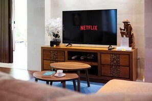 A room with a television displaying 'NETFLIX,' a wooden cabinet with shoes, a small round table with a book and a cup, and a statue.