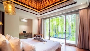 A modern bedroom with a large bed, a desk, and a view of greenery through the window.