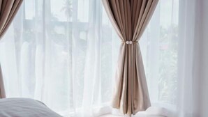 Quarto com cama, cortinas leves e vista para palmeiras lá fora.
